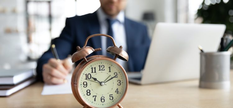 Focus on clock standing on table with busy young businessman entrepreneur in formal wear on background, focused millennial male manager involved in casual working process, time management concept.